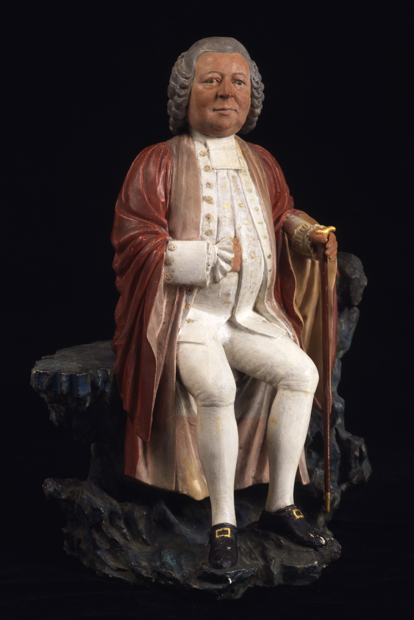 Sculpture of a white man in red robes, with a curly grey wig, seated on a crag of rock, holding a cane.
