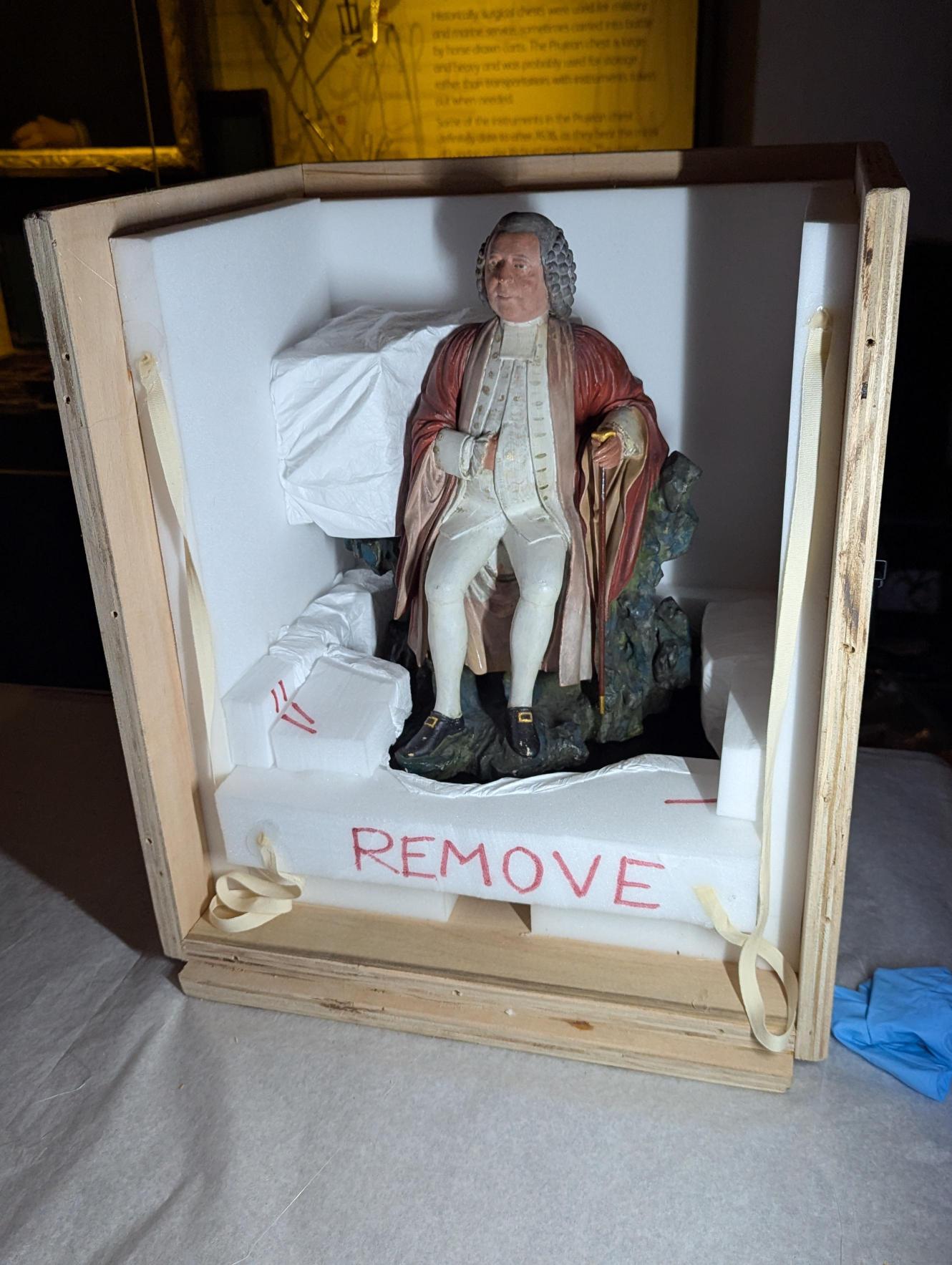Statuette of a man in red robes seated on a rock, packed within a wooden case, open on two sides so that the sculpture is completely visible.