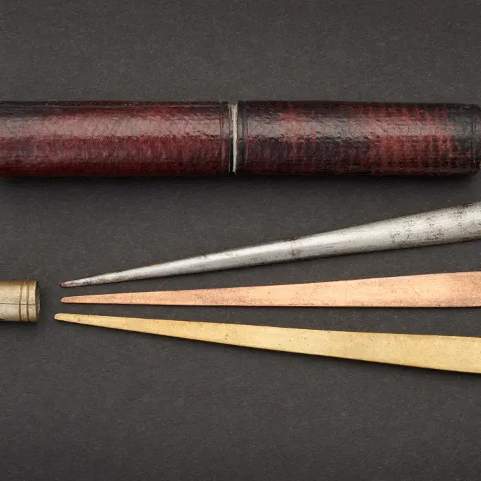 A cylindrical dark red leather case, above three rod in different metal with a tapering end and a rounded end, all three pointing at a metal collar.